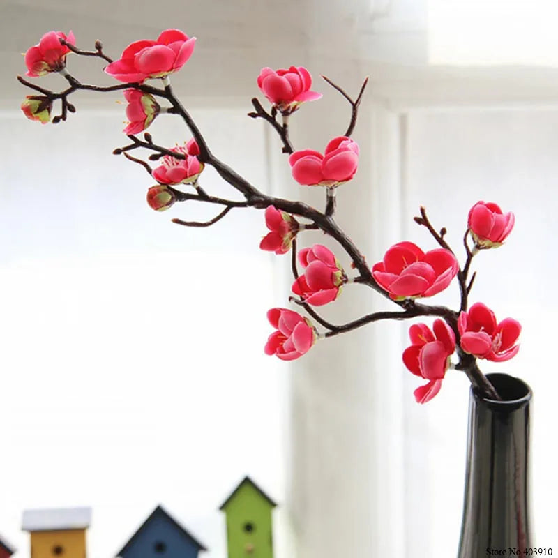 Pink artificial silk cherry blossom branches with vibrant red flowers displayed in a vase against a bright background.