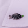 Silver enamel pin shaped like a fish with a green accent, resting on white fabric.