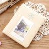 A beige compact photo album with a clear window displaying an instant film photo, resting on a wooden surface next to lace.