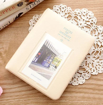 A beige compact photo album with a clear window displaying an instant film photo, resting on a wooden surface next to lace.