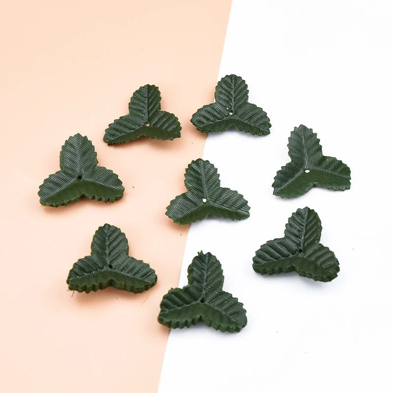 A collection of small, dark green silk artificial leaves scattered on a light pink and white background.