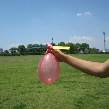 A hand holds a pink balloon with yellow streamers against a bright blue sky and green field.
