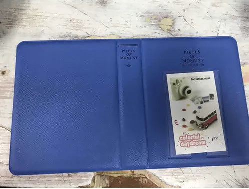 A blue compact photo album titled Pieces of Moment for instant film is shown on a wooden surface.