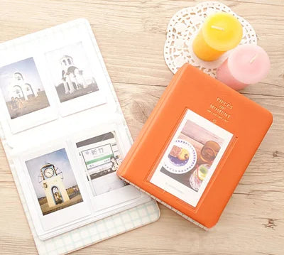 An orange compact photo album with a clear window displaying instant film photos and a yellow candle.