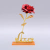 A gold foil artificial rose with a stem and leaves stands on a base engraved with the word love.