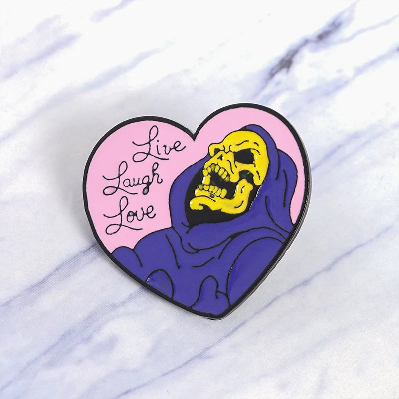 A heart-shaped enamel pin featuring a yellow skull wearing a purple hood with the text Live Laugh Love.