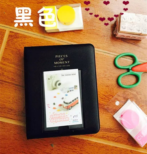 A black compact photo album for instant film is displayed on a wooden surface next to yellow and red heart decorations.