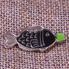 Silver enamel pin shaped like a fish with black body and green fins on a woven fabric background.