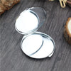 A silver mini double-sided folding makeup mirror is shown open on a dark wooden surface.