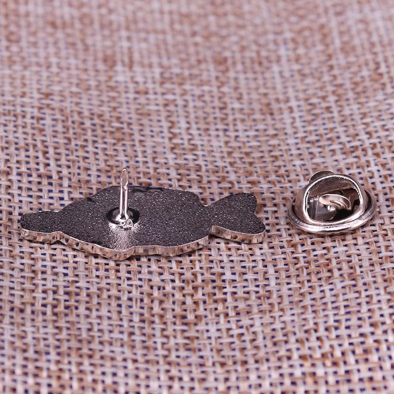 A silver enamel pin shaped like a fish resting on a textured fabric surface.