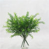 A vibrant green artificial fern and leaf sprig arrangement displayed in a clear glass vase.