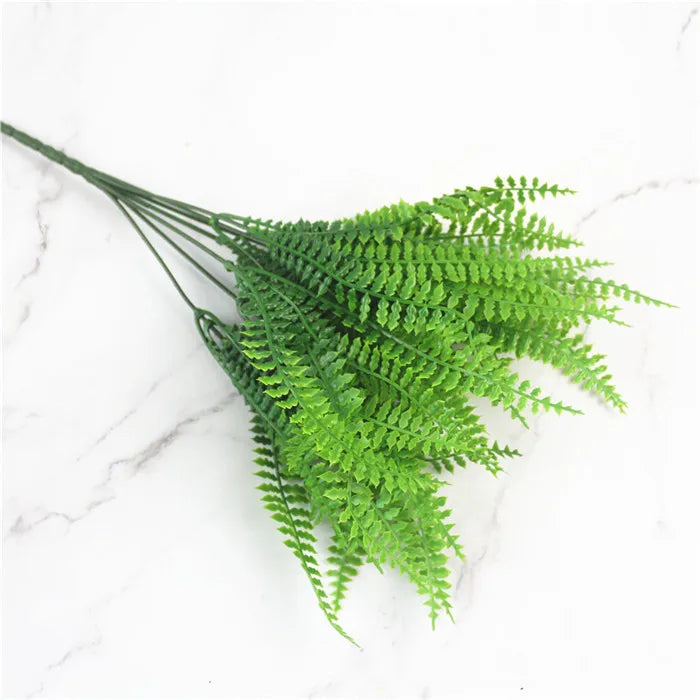 Green artificial fern and leaf sprigs arranged on a white marble surface.