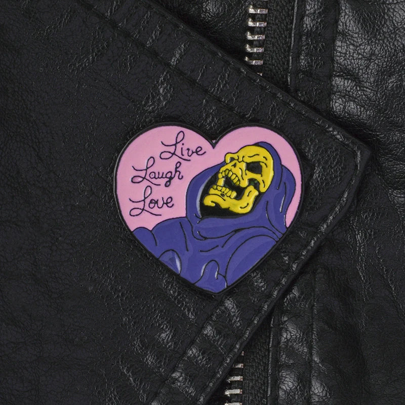 A heart-shaped enamel pin featuring a yellow skull design with the text Live Laugh Love on a purple background.