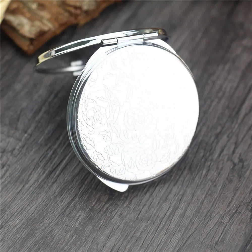 A small, round, silver-framed folding makeup mirror with an intricate design resting on a dark wooden surface.