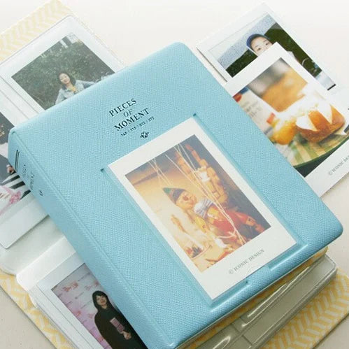 A light blue compact photo album with a textured cover lying next to several instant film photos.