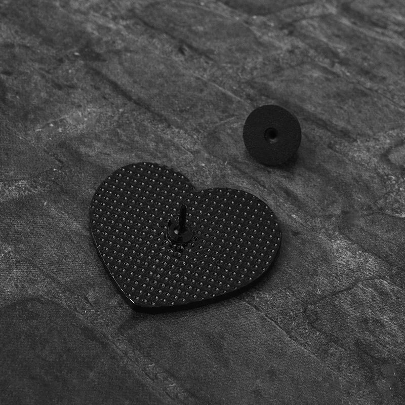A black heart-shaped enamel pin rests on a dark, textured surface next to a small black button.