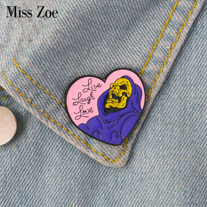 A heart-shaped enamel pin featuring a skull with purple shading and the text Live Laugh Love pinned on denim fabric.