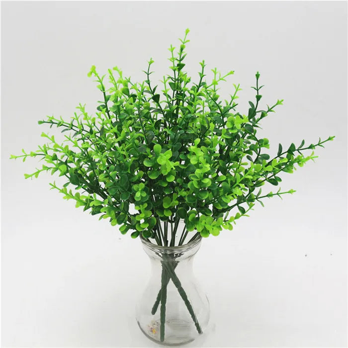 A bouquet of bright green artificial fern and leaf sprigs in a clear glass vase.