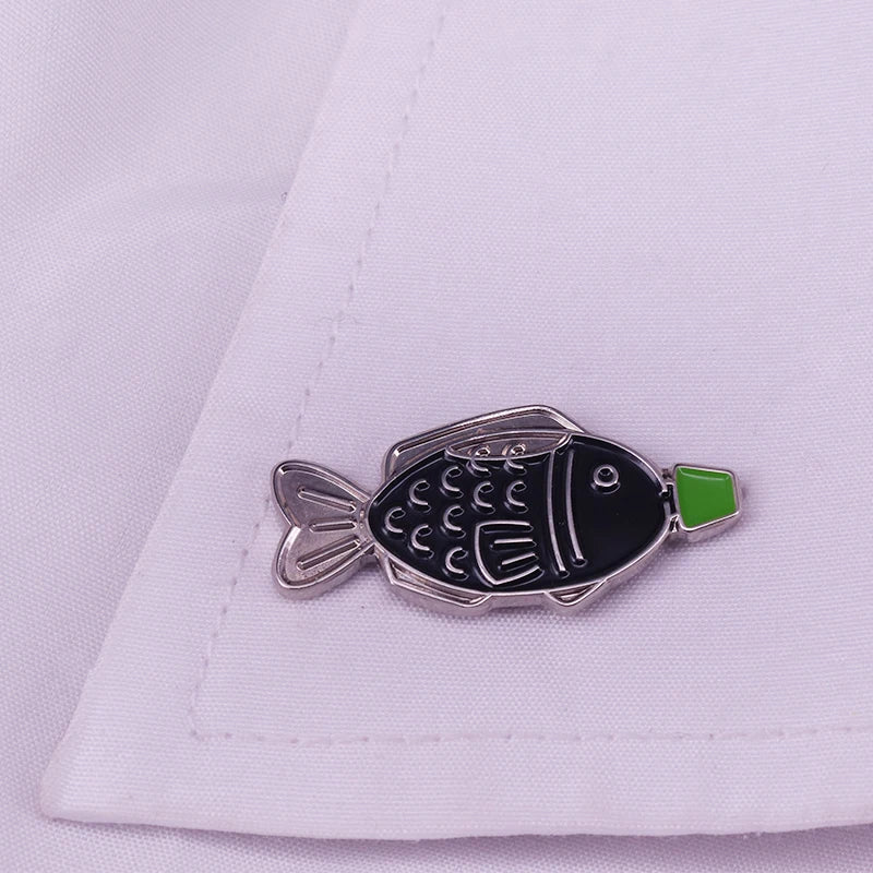 Silver enamel pin shaped like a fish with a green accent, resting on white fabric.