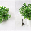 A close-up of vibrant green artificial fern and leaf sprigs with small white flowers displayed in a white vase.