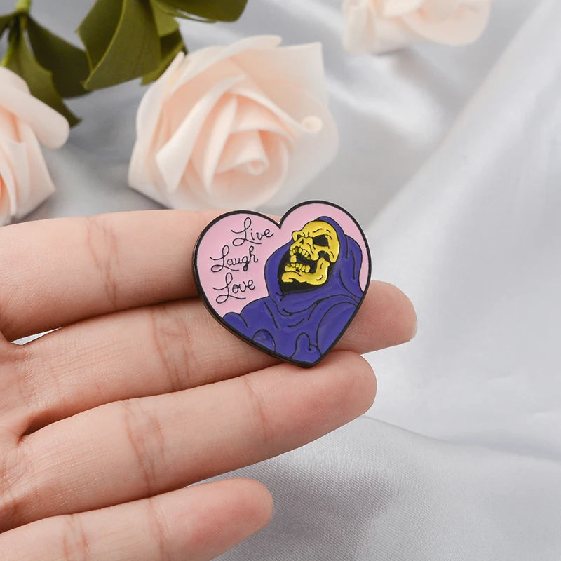 A hand holds a heart-shaped enamel pin featuring a yellow skull with the text Live Laugh Love on it.