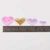 Three heart-shaped confetti pieces in pink, gold, and lavender shades displayed on a ruler.