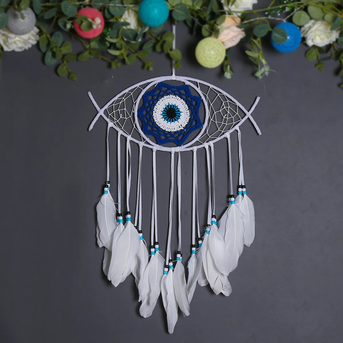 Handmade boho evil eye dream catcher with white feathers and blue mandala design hanging from green foliage