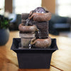 A stacked, rustic brown tabletop water fountain with a clear sphere at the top, set on a dark square dish.