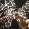 A group of people are holding and releasing clear, sparkling party balloons at an outdoor event.