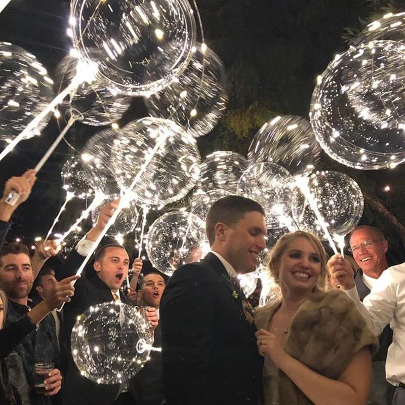 A group of people are holding and releasing clear, sparkling party balloons at an outdoor event.