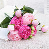 A bouquet of pink and magenta silk rose and peony artificial flowers with green leaves on a white surface.