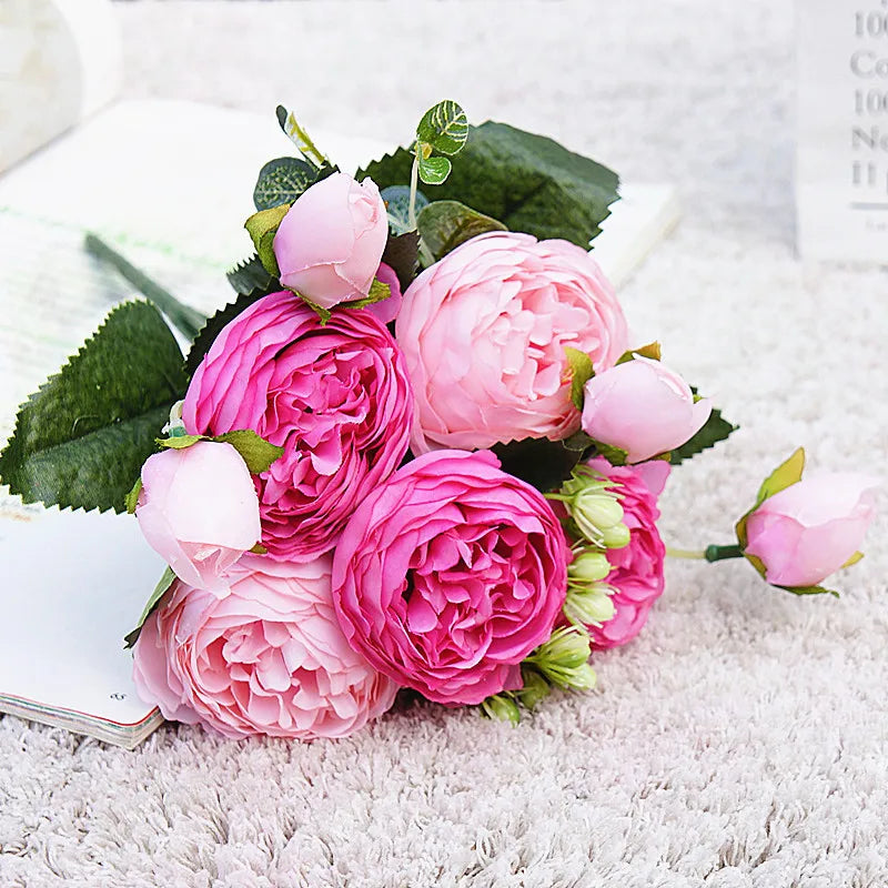 A bouquet of pink and magenta silk rose and peony artificial flowers with green leaves on a white surface.