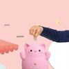 A pink cartoon cat piggy bank is being held by a hand against a light pink background.