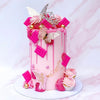 A pink cake decorated with pink and silver butterfly toppers and dripping icing on a white cake stand.