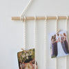 A macrame photo display featuring multiple photos hanging from a wooden bar on a white wall.