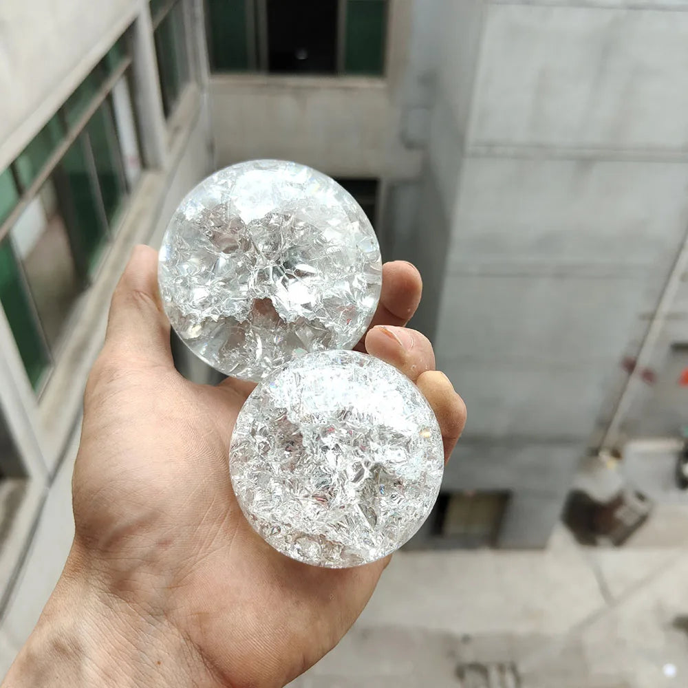 A hand holds two clear crystal ball water feature ornaments against a blurred building background.