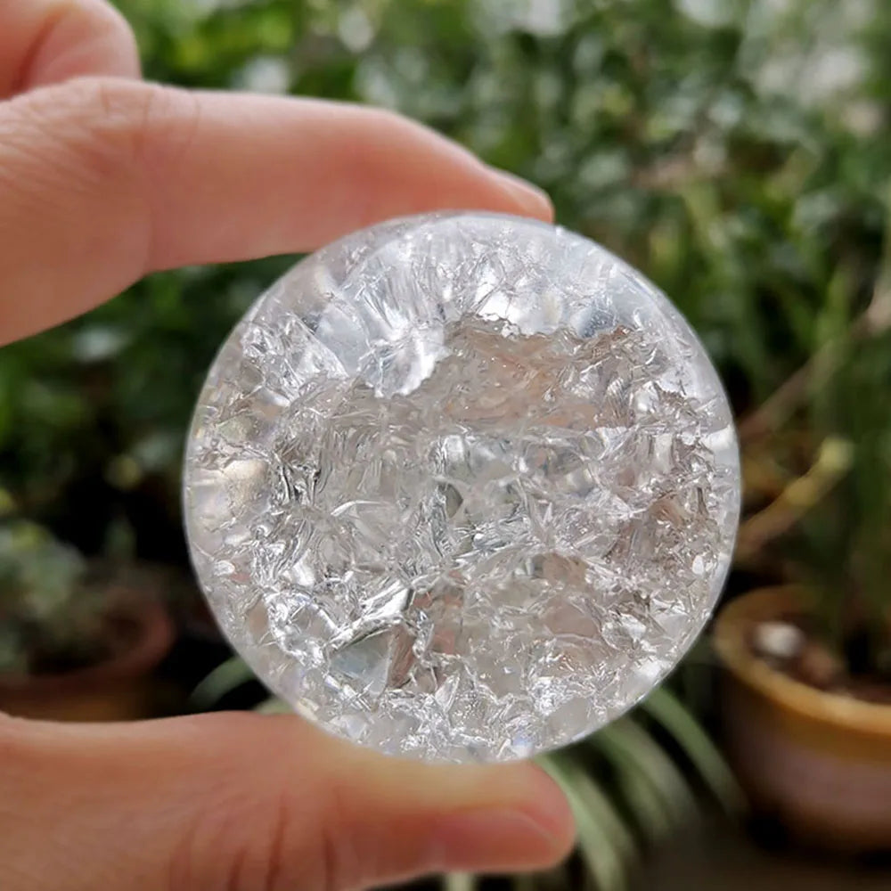 A hand holds a clear, round crystal ball water feature ornament against a blurred green background.