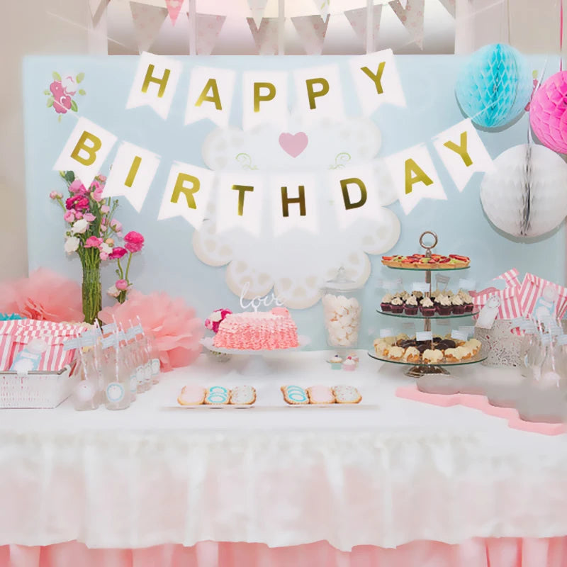 Decoración de pancarta de cumpleaños azul claro con letras doradas y nubes, sobre una mesa con dulces rosas y blancos.