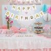 Decoración de pancarta de cumpleaños azul claro con letras doradas y nubes, sobre una mesa con dulces rosa y blanco.