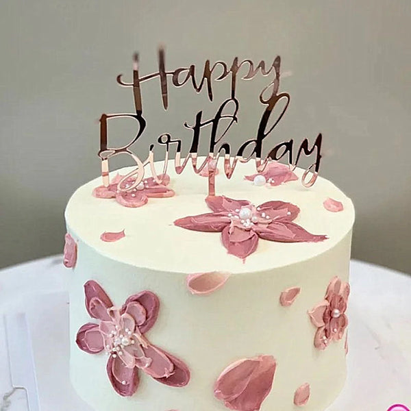 Pastel de cumpleaños blanco decorado con detalles florales rosas y una figura acrílica dorada que dice Feliz Cumpleaños