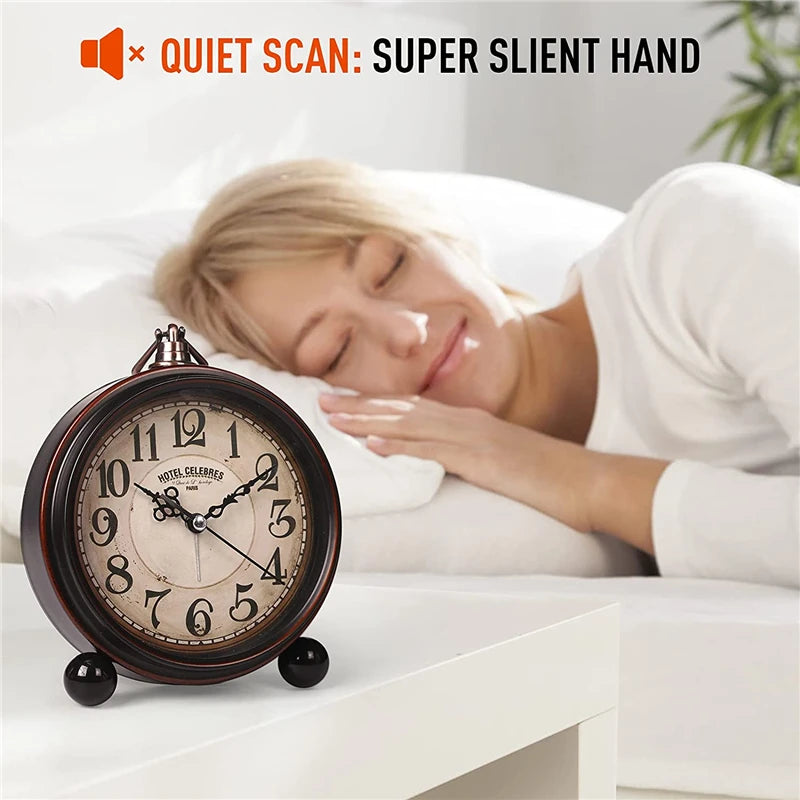 A vintage metal battery alarm clock is placed on a white surface next to a woman sleeping in bed.