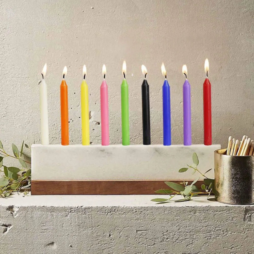 A row of 100 colored mini taper candles in various shades of orange, yellow, pink, green, black, and blue displayed on a whit