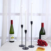 A set of tall white candles in black holders displayed on a white table with a bottle of red wine and some food.