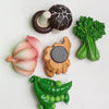 A set of realistic fruit and vegetable fridge magnets including a dark plum, a pink onion, a green broccoli floret, and a gre