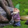 A miniature resin gnome statue with a green hat and white beard sitting on green grass next to a large rock.