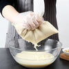 A hand wearing a white glove is squeezing a clear nylon filter bag filled with light-colored liquid into a glass bowl.
