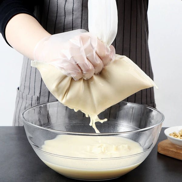 A hand wearing a white glove is squeezing a clear nylon filter bag filled with light-colored liquid into a glass bowl.