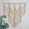 A cream-colored macrame wall hanging tapestry with fringe and tassels is displayed against a white wall.