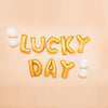 Gold foil letter balloons spelling LUCKY DAY with white balloons on a light background