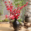 Vibrant red artificial silk cherry blossom branches arranged in a dark vase with greenery and natural wood elements.